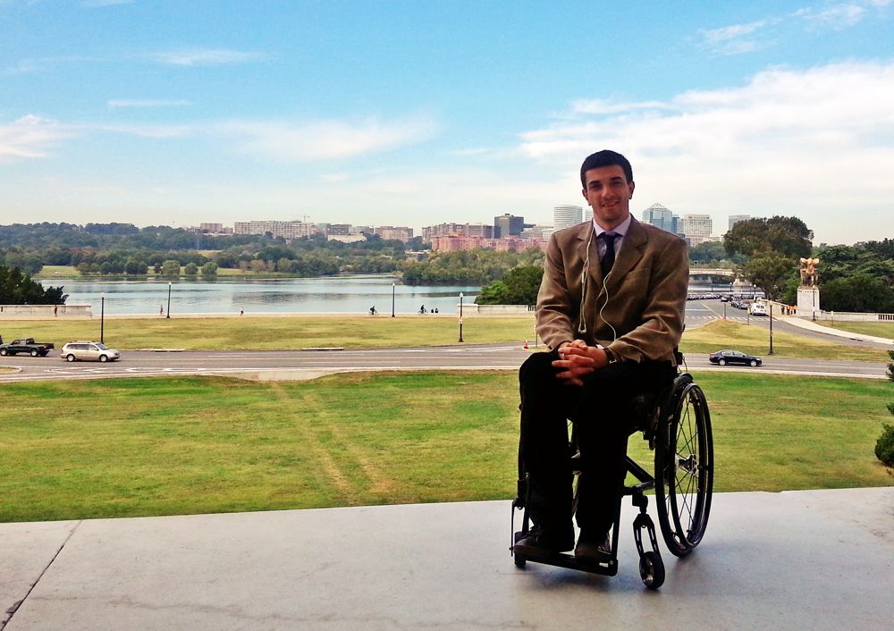 A gentleman in a light coat and bark pants sits in awheelchair at the egre of a wide lawn overlooking the water