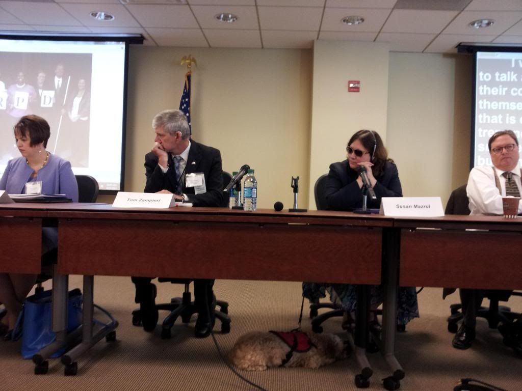 Two men and two women sit at a panel table, one with a service dog on the floor beneath