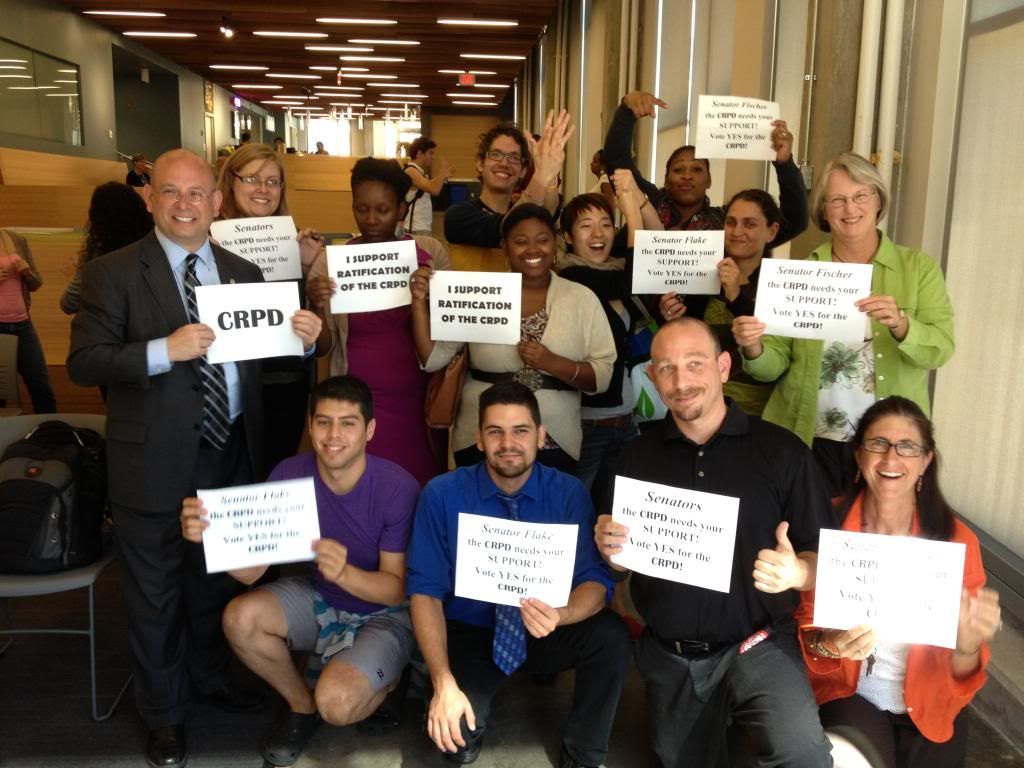 A group of 13 people of all ages joyfully hold up signs supporting the CRPD