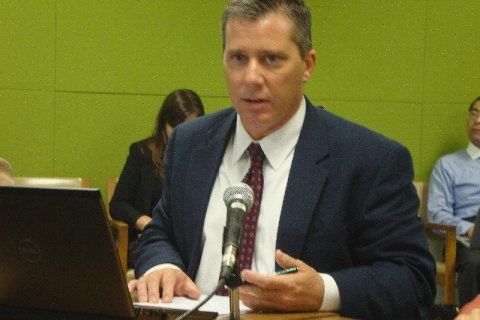 A man with slightly greying hair sits in front of a green background with a microphone brfore him and waering a business suit.